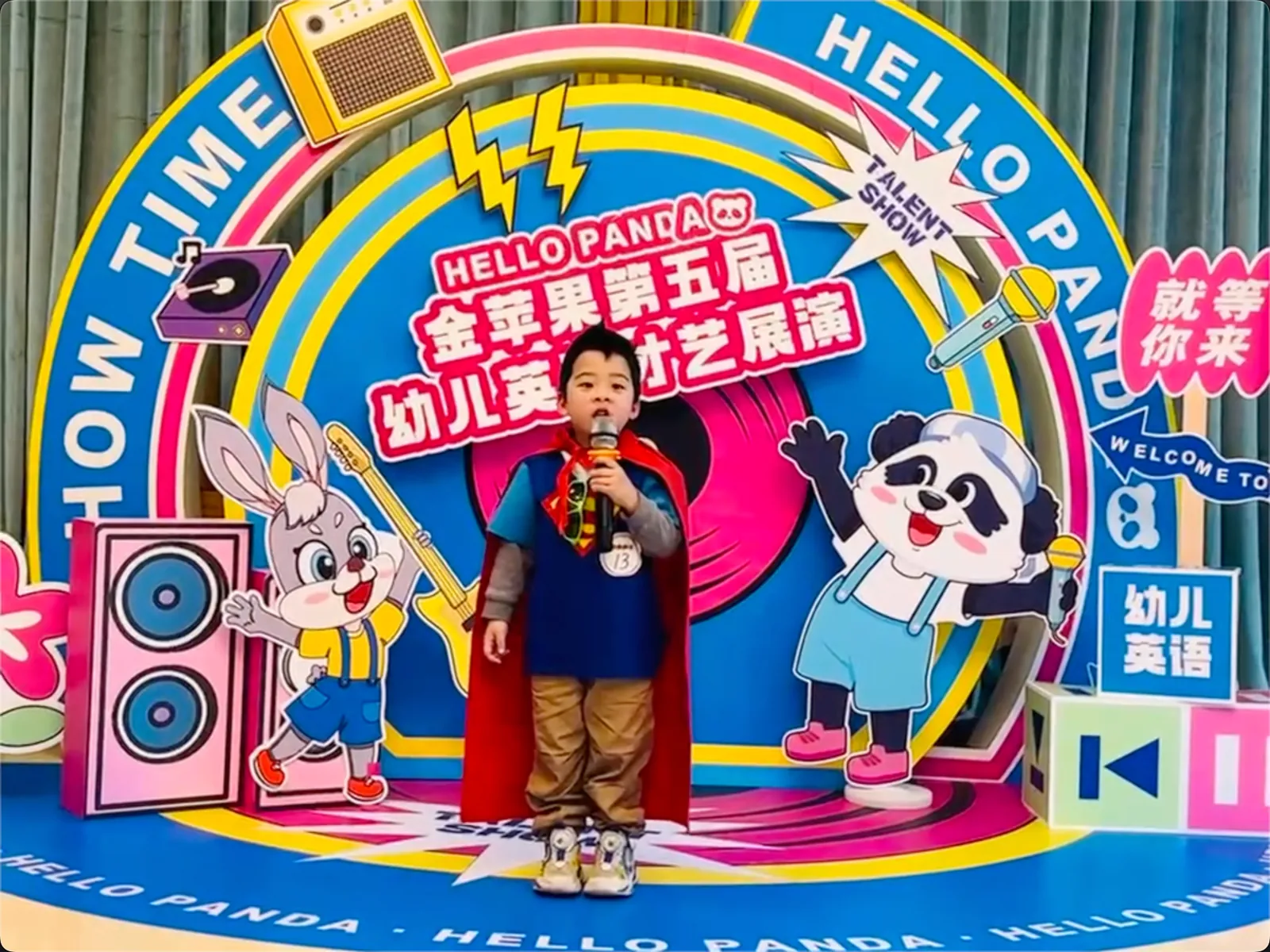 派派穿着披风在英语才艺展演背景前拿着话筒