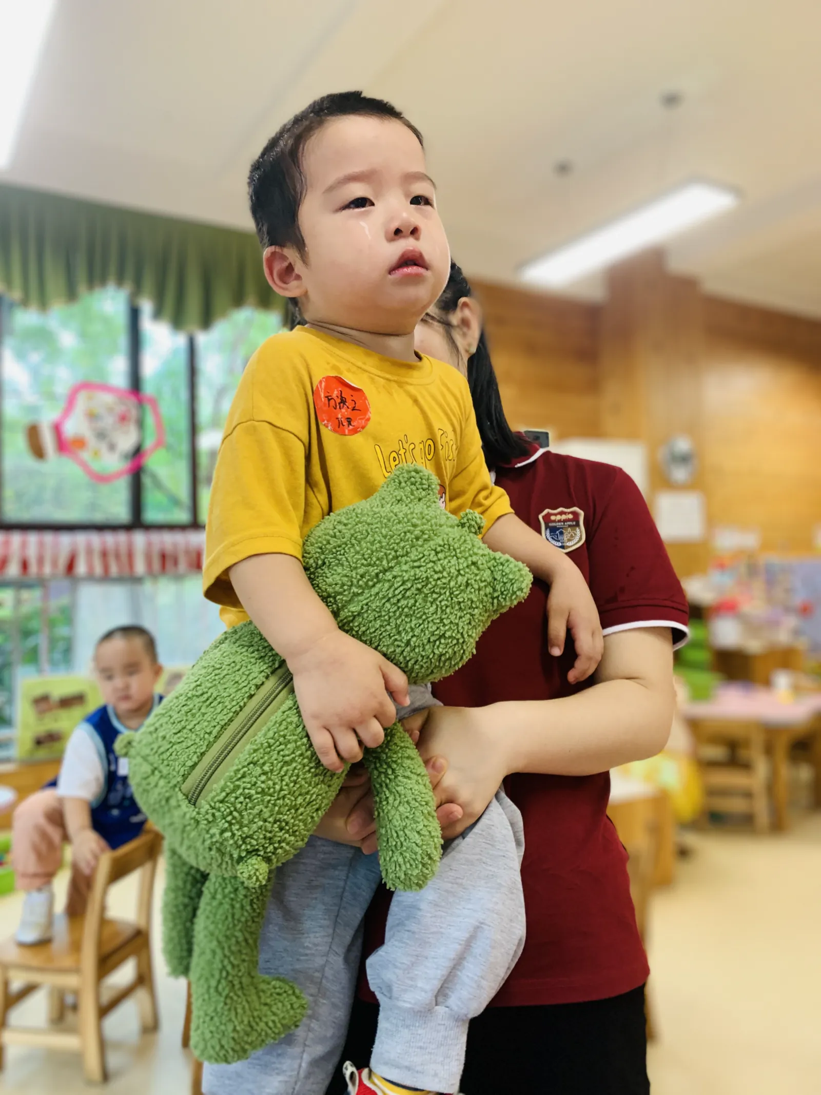 刚入园的派派抱着绿色玩偶，被老师抱在怀里安抚